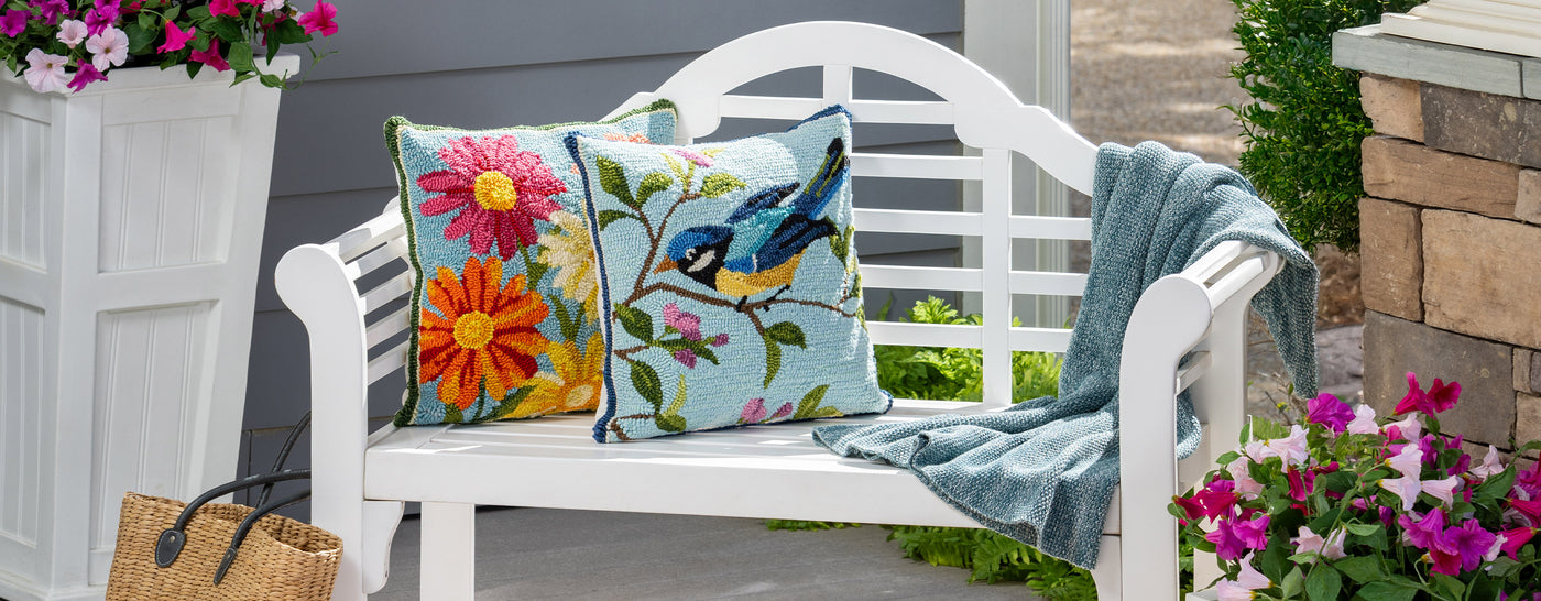 white outdoor Lutyens bench with colorful hooked pillows and a throw surrounded by flowers on a outdoor patio