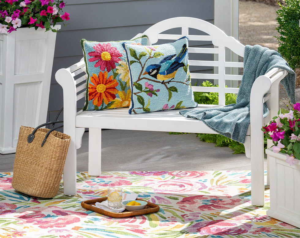 white outdoor Lutyens bench with colorful hooked pillows and a throw surrounded by flowers on a outdoor patio